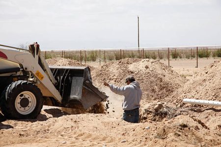 A young hispanic man signals to a backhoe operator while stones fall into the drain field of a new septic system.